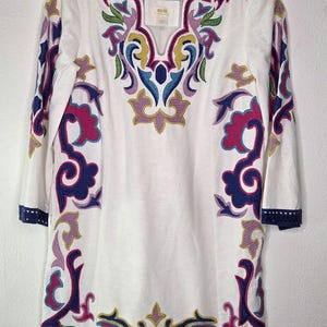 May include: White tunic dress with colorful embroidered floral patterns in pink, blue, yellow, and green. The dress has three-quarter sleeves and a navy blue trim along the neckline, sleeves, and hem. The brand name is visible on the neckline.