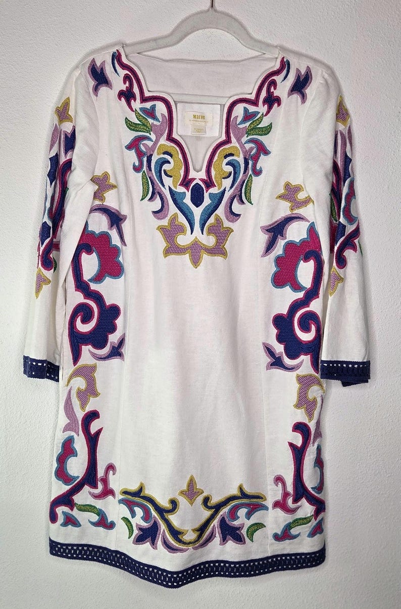 May include: White tunic dress with colorful embroidered floral patterns in pink, blue, yellow, and green. The dress has three-quarter sleeves and a navy blue trim along the neckline, sleeves, and hem. The brand name is visible on the neckline.