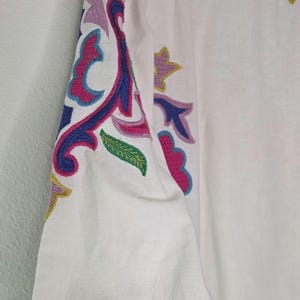 May include: White long-sleeved top with colorful embroidered floral and abstract designs in pink, blue, purple, and yellow. The sleeve cuff has a navy blue crocheted trim. The top is made of a lightweight fabric.