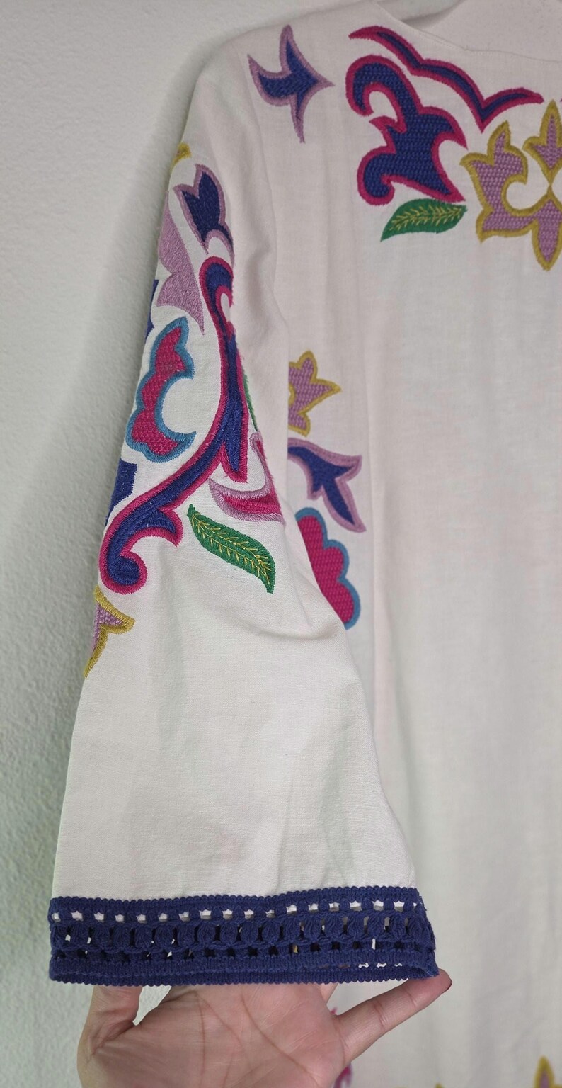 May include: White long-sleeved top with colorful embroidered floral and abstract designs in pink, blue, purple, and yellow. The sleeve cuff has a navy blue crocheted trim. The top is made of a lightweight fabric.