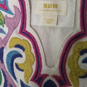 May include: White top with colorful embroidered floral design in pink, blue, yellow, and purple. The top features a V-neckline and a label that reads "Maeve by Anthropologie" and "X-Small Made in India."