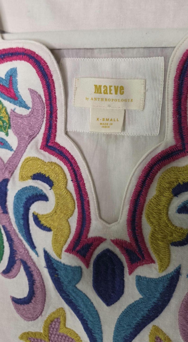 May include: White top with colorful embroidered floral design in pink, blue, yellow, and purple. The top features a V-neckline and a label that reads "Maeve by Anthropologie" and "X-Small Made in India."