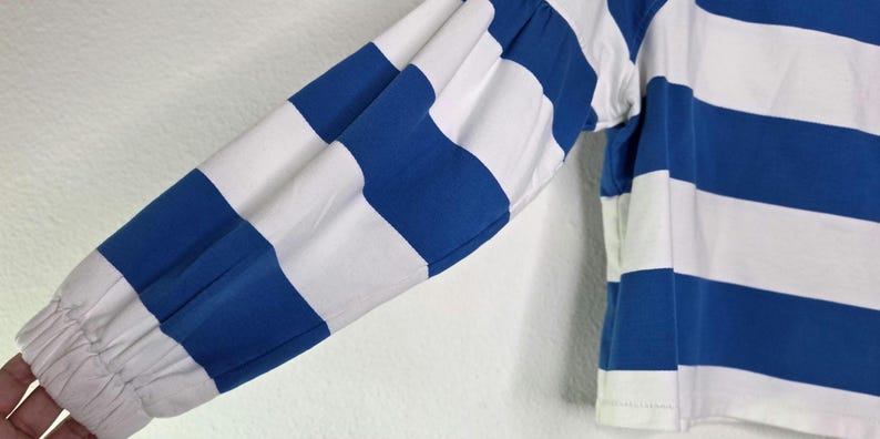 May include: Close-up of a long-sleeved shirt with blue and white horizontal stripes. The sleeve is gathered at the wrist with elastic. The shirt is made of a soft, comfortable material.