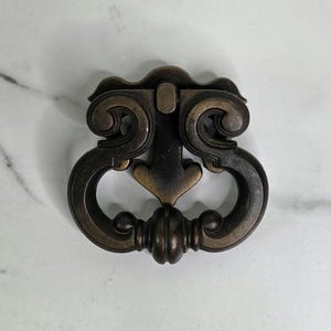 May include: An antique bronze-toned drawer pull with an ornate design. The pull features a decorative top with scrolling details and a central arrow-shaped element. The handle is a curved, semi-circular shape with a ribbed, rounded base. The hardware is set against a white marble surface.