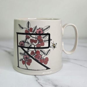 Puede incluir: Taza de cer&aacute;mica color crema con una letra negra "R" con un dise&ntilde;o floral de bayas rojas, flores rosas y hojas verdes. Tambi&eacute;n se representa una peque&ntilde;a abeja. La taza tiene un asa curva.