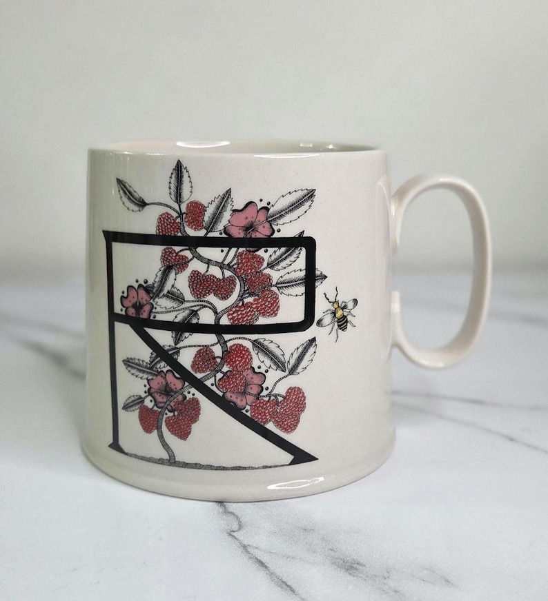 Puede incluir: Taza de cer&aacute;mica color crema con una letra negra "R" con un dise&ntilde;o floral de bayas rojas, flores rosas y hojas verdes. Tambi&eacute;n se representa una peque&ntilde;a abeja. La taza tiene un asa curva.
