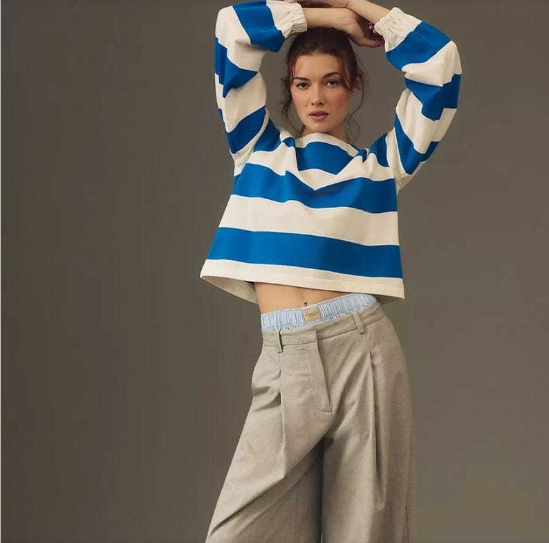 May include: A cropped, long-sleeved top with horizontal blue and white stripes. The model is wearing light gray pleated trousers. The top has elastic cuffs and a round neckline. The model is posing with their arms above their head.