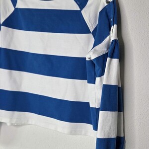 May include: A blue and white striped long-sleeve shirt with a round neckline and button details on the shoulder. The shirt is hanging on a white hanger against a white wall.