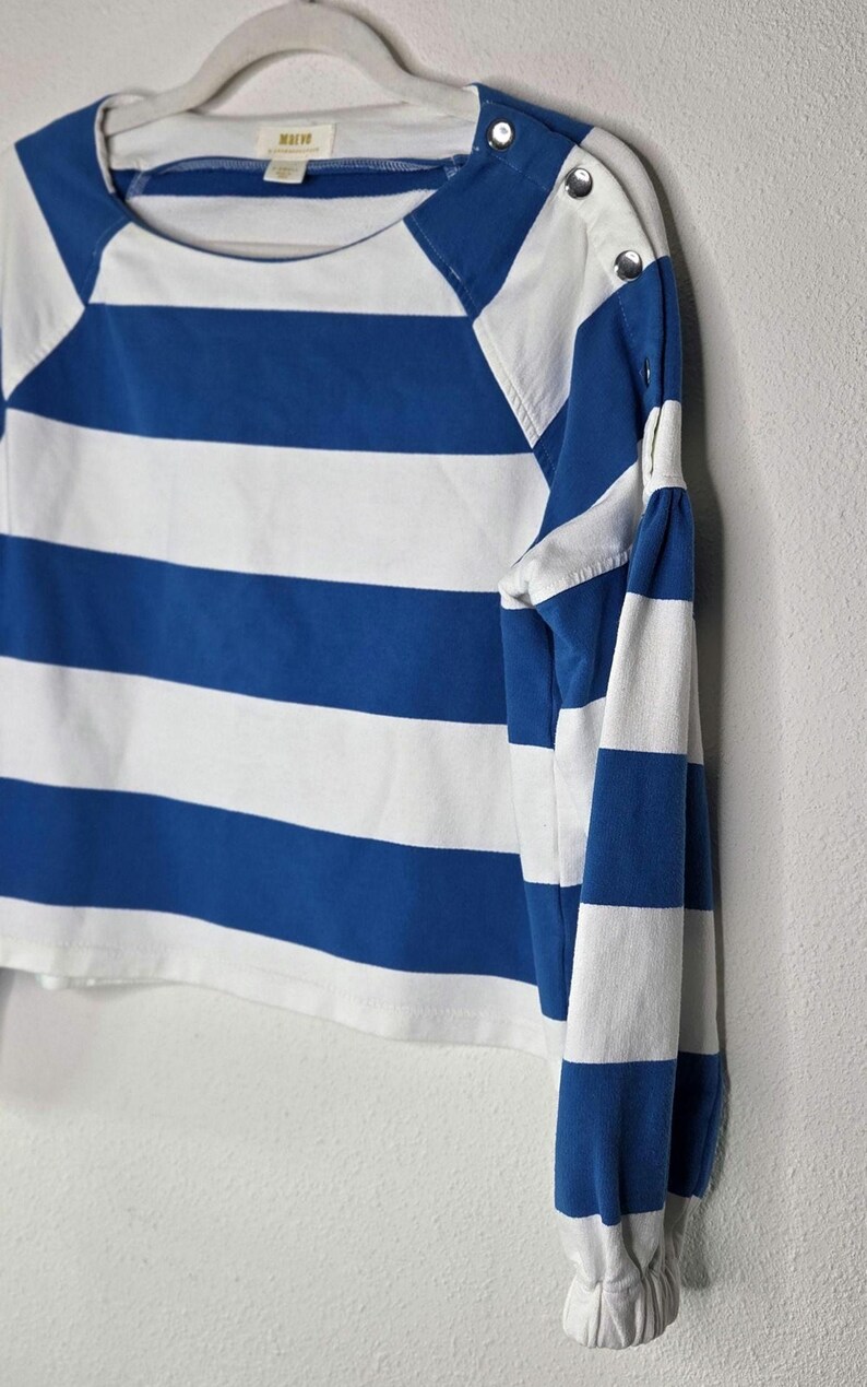 May include: A blue and white striped long-sleeve shirt with a round neckline and button details on the shoulder. The shirt is hanging on a white hanger against a white wall.