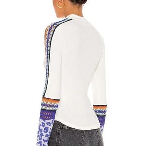 May include: White long-sleeve top with patterned sleeves. The sleeves feature a mix of colors including blue, orange, and purple. The top is paired with dark gray jeans. The model has dark hair and gold hoop earrings.