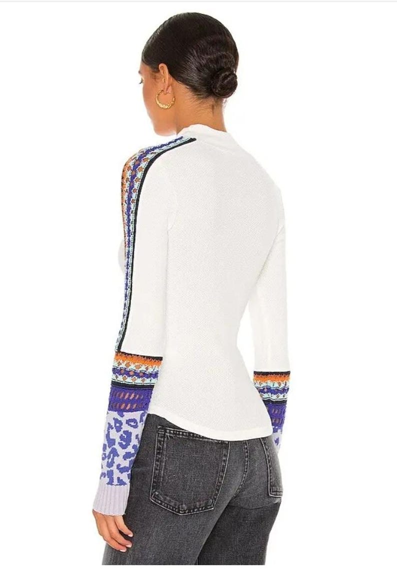 May include: White long-sleeve top with patterned sleeves. The sleeves feature a mix of colors including blue, orange, and purple. The top is paired with dark gray jeans. The model has dark hair and gold hoop earrings.