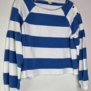 May include: A blue and white striped long-sleeve shirt with a round neckline and button details on the shoulder. The sleeves are gathered at the wrist. The shirt is hanging on a white hanger.