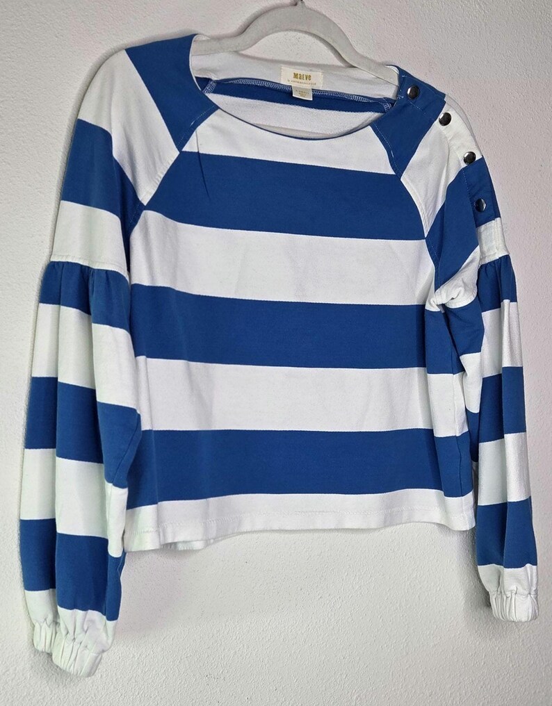 May include: A blue and white striped long-sleeve shirt with a round neckline and button details on the shoulder. The sleeves are gathered at the wrist. The shirt is hanging on a white hanger.