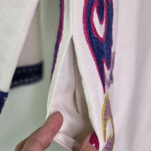 May include: Close-up of a white garment with colorful embroidered floral designs. The embroidery features shades of blue, pink, purple, and yellow. The garment has a pocket and a slit on the side.