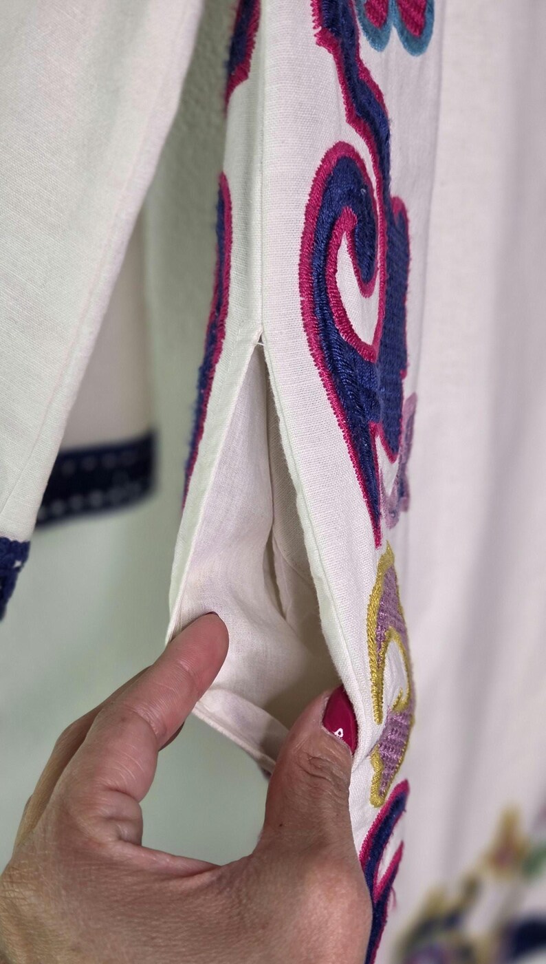 May include: Close-up of a white garment with colorful embroidered floral designs. The embroidery features shades of blue, pink, purple, and yellow. The garment has a pocket and a slit on the side.