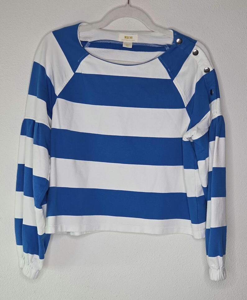 May include: A blue and white striped long-sleeve shirt. The shirt features wide horizontal stripes and a round neckline. The sleeves have a gathered, puffy design at the elbow and are cuffed at the wrist. The shoulder has three decorative buttons. The brand name "Maive" is visible.