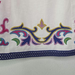 May include: White fabric with colorful embroidered floral designs in pink, blue, green, and yellow. The bottom edge features a navy blue trim with a decorative pattern. The embroidery is detailed and vibrant, creating a decorative border.