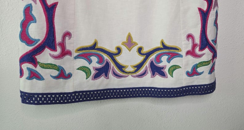 May include: White fabric with colorful embroidered floral designs in pink, blue, green, and yellow. The bottom edge features a navy blue trim with a decorative pattern. The embroidery is detailed and vibrant, creating a decorative border.