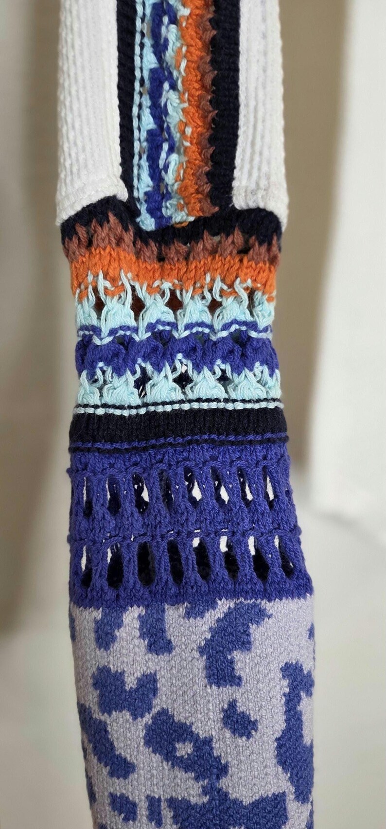 May include: Close-up of a colorful, patterned knitted sleeve. The design features a mix of white, orange, blue, and black sections. The lower portion has a blue and gray animal print.
