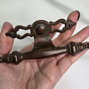 May include: Antique bronze drawer pull with ornate detailing. The pull features a central circular element and decorative scrollwork on the top and ends. The bronze has a weathered patina, adding to its vintage charm.