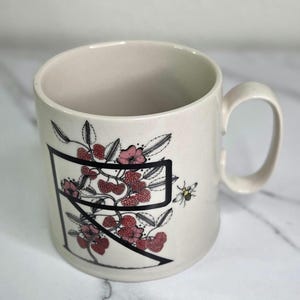 Puede incluir: Taza de cer&aacute;mica color crema con una letra "R" negra y un dise&ntilde;o floral de bayas rojas, flores rosas y hojas verdes. Tambi&eacute;n se representa una peque&ntilde;a abeja. La taza tiene un asa curva.