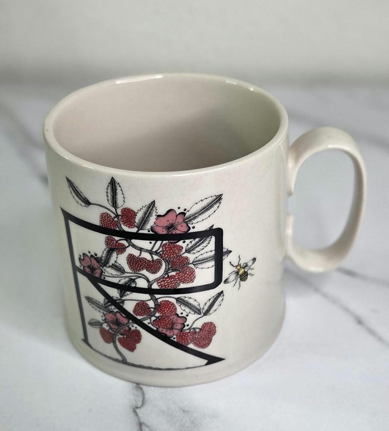 Puede incluir: Taza de cer&aacute;mica color crema con una letra "R" negra y un dise&ntilde;o floral de bayas rojas, flores rosas y hojas verdes. Tambi&eacute;n se representa una peque&ntilde;a abeja. La taza tiene un asa curva.