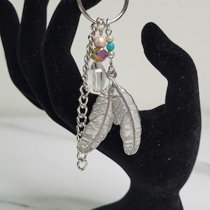 May include: Silver-toned keychain with feather charms and colorful beads. The keychain features two detailed feather pendants, a chain, and a ring. The beads include pink, turquoise, and iridescent accents. A stylish accessory for keys or bags.