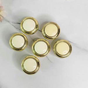 May include: Seven round buttons with a cream-colored center and a gold-colored rim. The buttons are arranged on a white and gray marble surface. The buttons are likely for sewing or crafts.