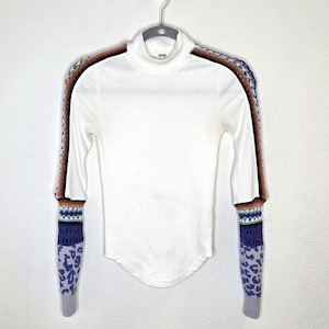 May include: White long-sleeved turtleneck shirt with patterned sleeves. The sleeves feature a mix of colors including brown, black, blue, and purple. The cuffs have a leopard print design.