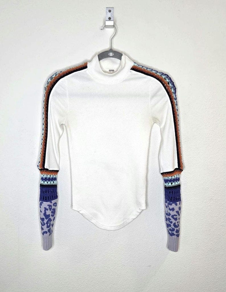 May include: White long-sleeved turtleneck shirt with patterned sleeves. The sleeves feature a mix of colors including brown, black, blue, and purple. The cuffs have a leopard print design.