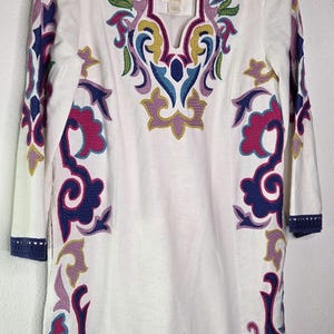 May include: White tunic dress with colorful embroidered floral patterns in purple, blue, pink, and yellow. The dress has three-quarter sleeves and a scalloped neckline. The hem and sleeve cuffs are trimmed with navy blue crochet.