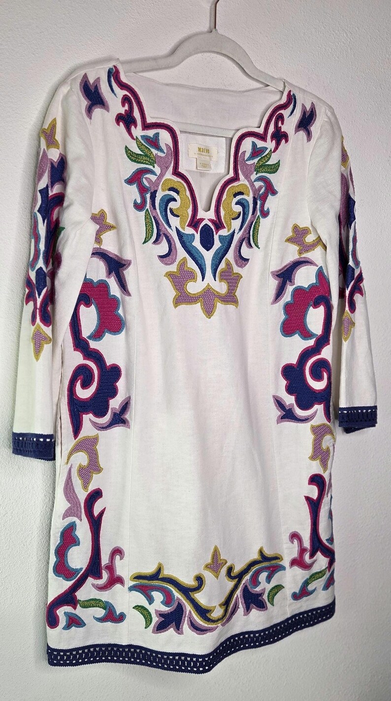 May include: White tunic dress with colorful embroidered floral patterns in purple, blue, pink, and yellow. The dress has three-quarter sleeves and a scalloped neckline. The hem and sleeve cuffs are trimmed with navy blue crochet.