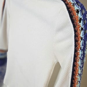May include: White long-sleeved shirt with a textured waffle knit. The sleeves feature a decorative crochet stripe in blue, orange, and teal. The shirt has a mock neck collar.