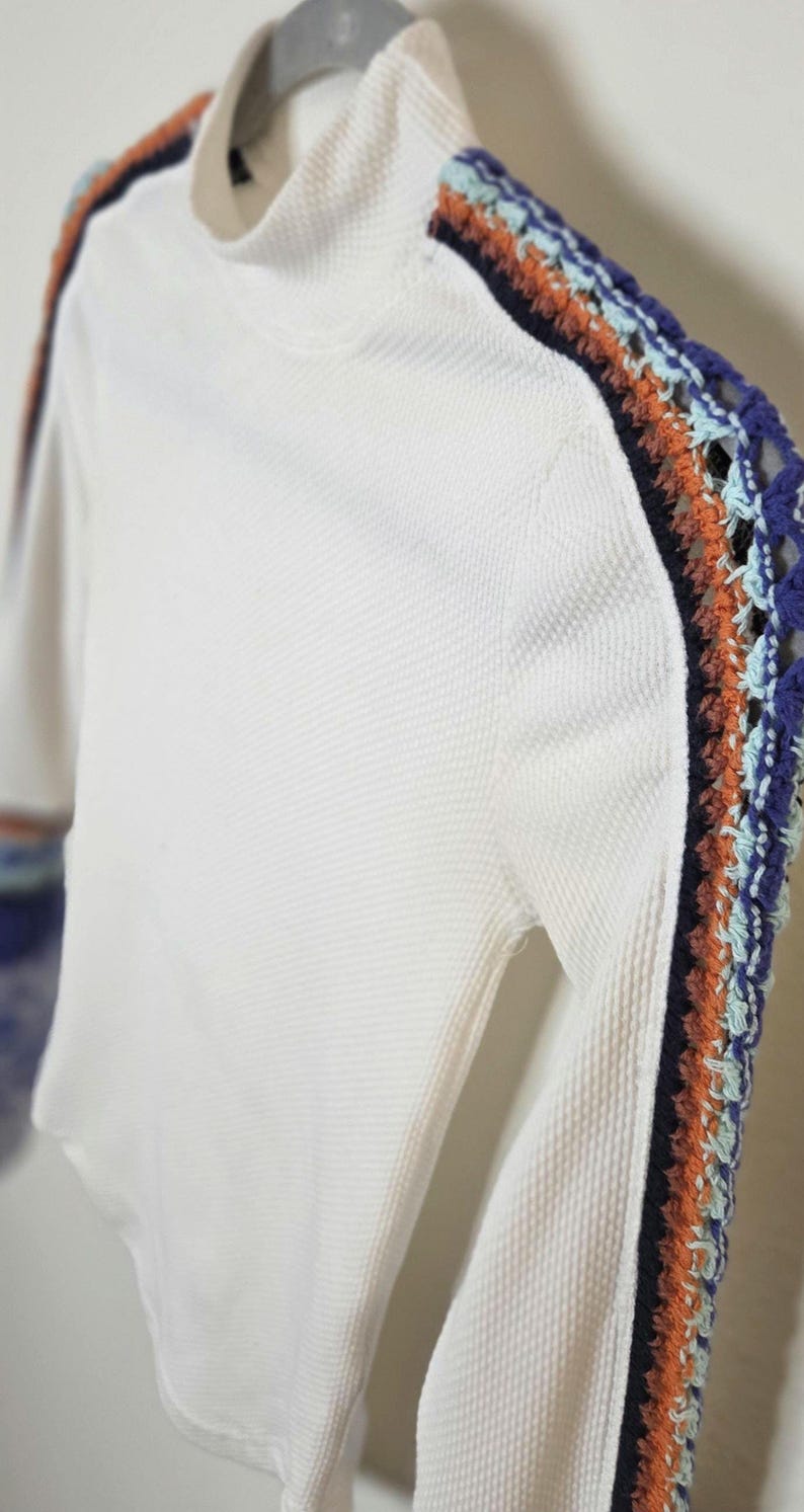 May include: White long-sleeved shirt with a textured waffle knit. The sleeves feature a decorative crochet stripe in blue, orange, and teal. The shirt has a mock neck collar.