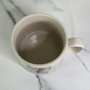 Puede incluir: Taza de cer&aacute;mica color crema con asa redondeada. El interior es de color beige claro y el exterior presenta un dise&ntilde;o sutil cerca de la base. La taza est&aacute; vac&iacute;a y se encuentra sobre una superficie de m&aacute;rmol blanco.