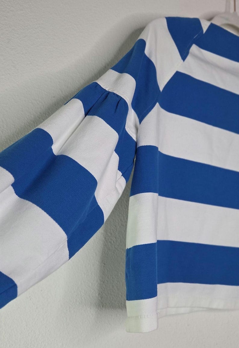 May include: A long-sleeved shirt with alternating horizontal stripes of white and blue. The sleeve is visible, showing the blue and white stripes. The shirt has a classic design, with a white collar and cuffs. The fabric appears to be a soft, breathable material.