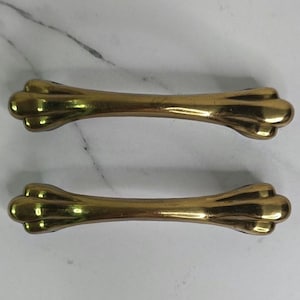 May include: Two antique brass drawer pulls. Each pull has a bone-shaped design with rounded ends. The pulls are a warm gold color and are set against a white and gray marble background. These pulls are likely for furniture.