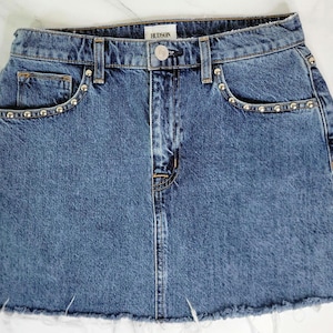 May include: A blue denim skirt with a raw hem. The skirt has silver stud accents along the front pockets and waistband. The brand name "HUDSON" is visible on the waistband.