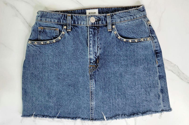 May include: A blue denim skirt with a raw hem. The skirt has silver stud accents along the front pockets and waistband. The brand name "HUDSON" is visible on the waistband.