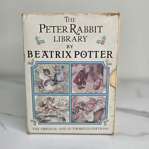 May include: A rectangular box set titled The Peter Rabbit Library by Beatrix Potter, featuring four distinct illustrations of anthropomorphic animals in muted colors. The white cover includes the text The Original and Authorized Editions.