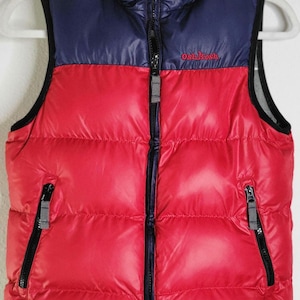May include: A red and navy blue puffer vest with a full zipper closure and two zippered pockets. The vest has a gray collar and the brand name "Oshkosh" embroidered on the chest. The vest is hanging on a white hanger.