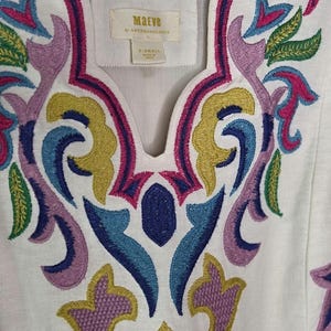 May include: White tunic top with colorful embroidered floral design. The embroidery features shades of pink, purple, blue, green, and yellow. The top has a scoop neckline and the brand name "Maeve" is visible on a tag.