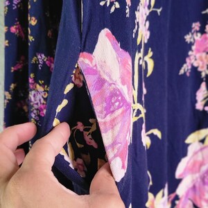 May include: Close-up of navy blue fabric with a floral print. The fabric displays large pink and purple flowers with yellow accents. A hand holds open a pocket, showing the inside of the garment. The overall theme is clothing.