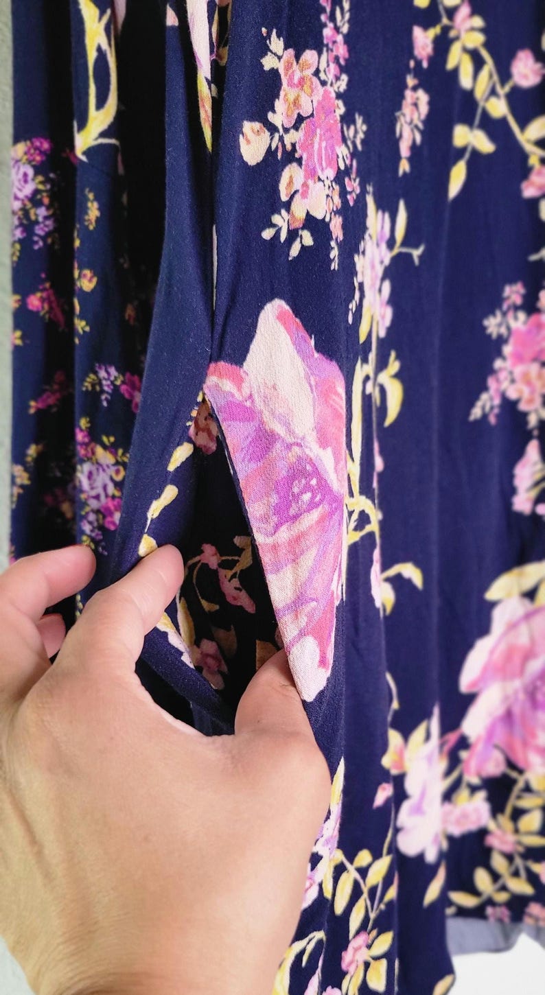 May include: Close-up of navy blue fabric with a floral print. The fabric displays large pink and purple flowers with yellow accents. A hand holds open a pocket, showing the inside of the garment. The overall theme is clothing.