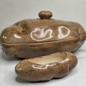 May include: A brown, potato-shaped ceramic lidded container with a small knob handle on top, accompanied by a smaller, open, potato-shaped ceramic dish with a smooth, cream-colored interior. Both pieces feature a glossy, speckled finish.