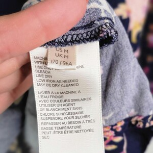 May include: A clothing label with care instructions in English and French. The label is white with black text and washing symbols. The fabric is navy blue with a floral pattern in pink and yellow.