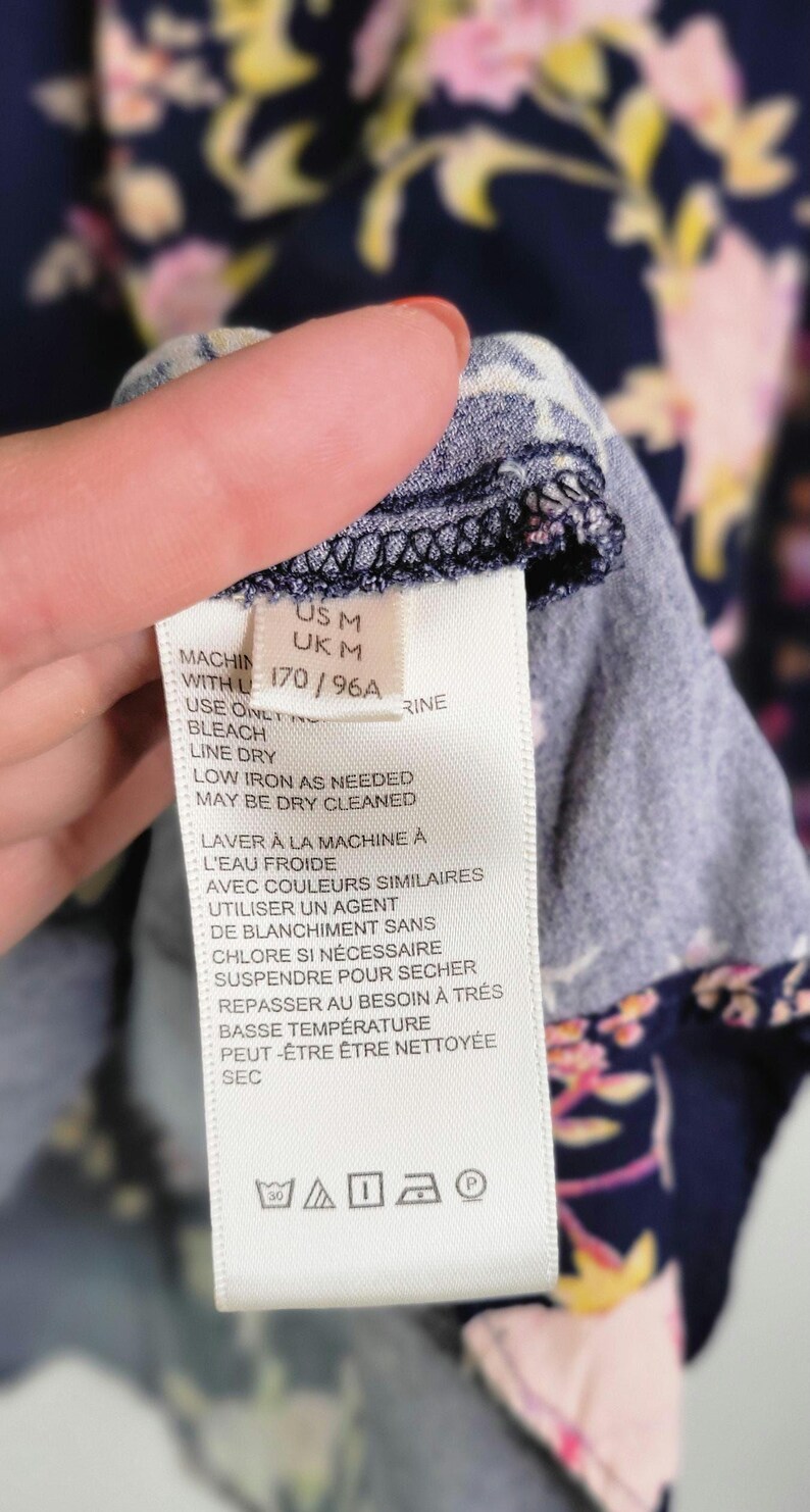May include: A clothing label with care instructions in English and French. The label is white with black text and washing symbols. The fabric is navy blue with a floral pattern in pink and yellow.