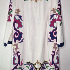 May include: White tunic dress with colorful embroidered floral patterns in purple, pink, green, and gold. The dress has long sleeves and a navy blue trim with a crochet design along the cuffs and hemline. The neckline is a simple scoop.