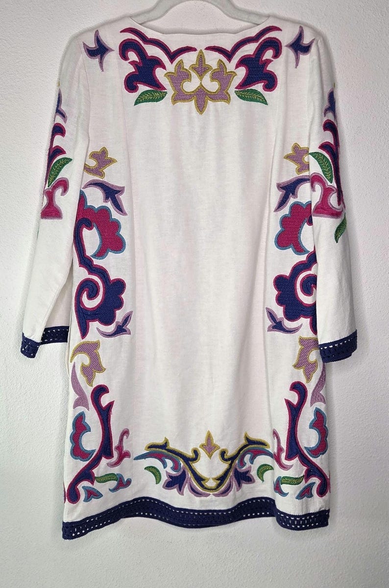 May include: White tunic dress with colorful embroidered floral patterns in purple, pink, green, and gold. The dress has long sleeves and a navy blue trim with a crochet design along the cuffs and hemline. The neckline is a simple scoop.