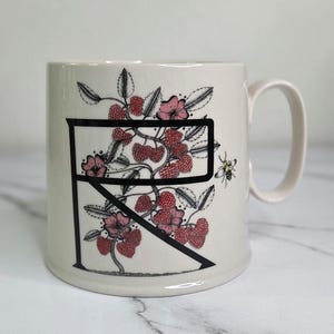 Puede incluir: Taza de cer&aacute;mica color crema con la letra negra &laquo;R&raquo;, adornada con bayas rojas, flores rosas y hojas verdes. Tambi&eacute;n se representa una peque&ntilde;a abeja. La taza tiene un asa curvada.